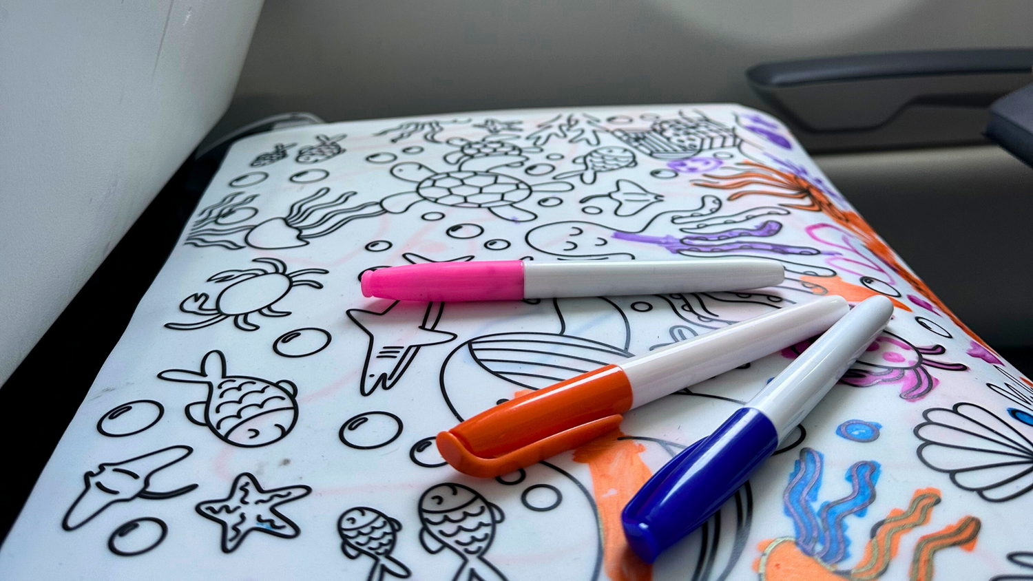 Under The Sea themed reusable coloring mat with colored dry erase markers, set on a airplane tray table